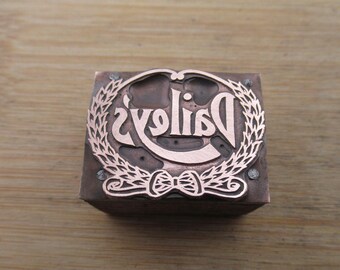 May include: A copper printing block featuring the word "Daisies" in a decorative font, framed by a laurel wreath and a bow. The square block rests on a wooden surface.
