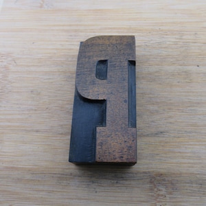 May include: A rectangular wooden letterpress block featuring the uppercase letter P in a bold, serif typeface. The block has a dark brown, weathered wood grain finish on the face, while the sides and recessed areas are painted a solid matte black.