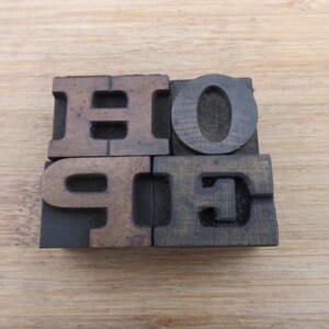 May include: Four individual dark brown wooden letterpress blocks arranged in a square formation. The blocks feature the raised letters H, O, P, and E, which together spell the word HOPE. The wood grain is visible on the surface of each letter.