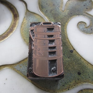 Op de afbeelding: Een vintage koperkleurige drukblok met een gedetailleerd ontwerp van een stapel van vijf ronde containers met een handvat aan de bovenkant.