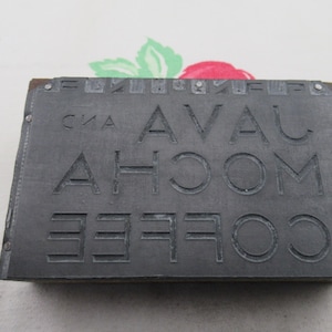 Op de afbeelding: Een grijs metalen drukblok met de woorden "AVA" en "ROOM" in reliëf. Het blok is licht beschadigd en heeft een paar roestplekken.