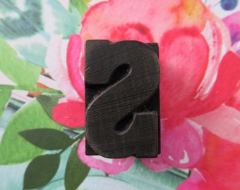 May include: A dark brown wooden letter block with the letter "S" carved into it. The block is sitting on a pink and green floral background.