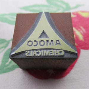 Puede incluir: Un bloque de madera marrón con un triángulo de metal en la parte superior. El triángulo es dorado y tiene el texto "AMO CO. CHEMICALS" impreso en él.