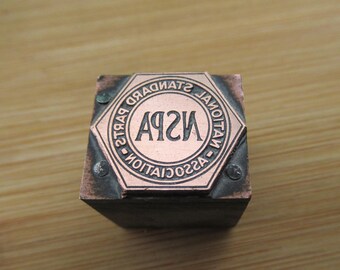 Może przedstawiać: Stempel metalowy o sześciokątnym kształcie z okrągłym wzorem. Wzór zawiera tekst "ASSOCIATION - STANDARD PARTS" otaczający tekst "ASPA" większą czcionką.