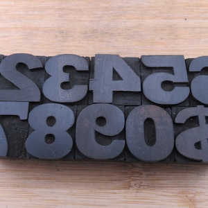 May include: A set of ten rectangular black letterpress blocks arranged in two rows. The top row features the numbers 1, 2, 3, 4, 5, and 6. The bottom row displays the numbers 7, 8, 9, 0, and a dollar sign symbol, all in a bold, sans-serif font.