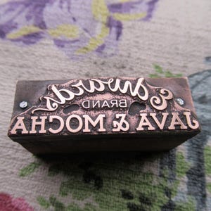 Peut inclure: Tampon rectangulaire de couleur cuivrée avec l'inscription "Brand Mocha" dans une police décorative et inversée. L'aspect est vintage, probablement pour le marquage ou l'artisanat. Le design comprend des volutes.