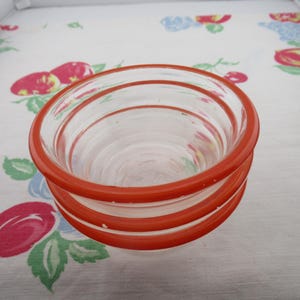 May include: A set of three clear glass bowls stacked vertically. Each bowl features a distinct, thick, bright orange rim around the top edge. The glass is transparent, showing the circular, ribbed texture of the bowls as they nest together.