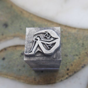 May include: A silver metal stamp with a design of a person sitting with their legs crossed and their arms wrapped around their legs.