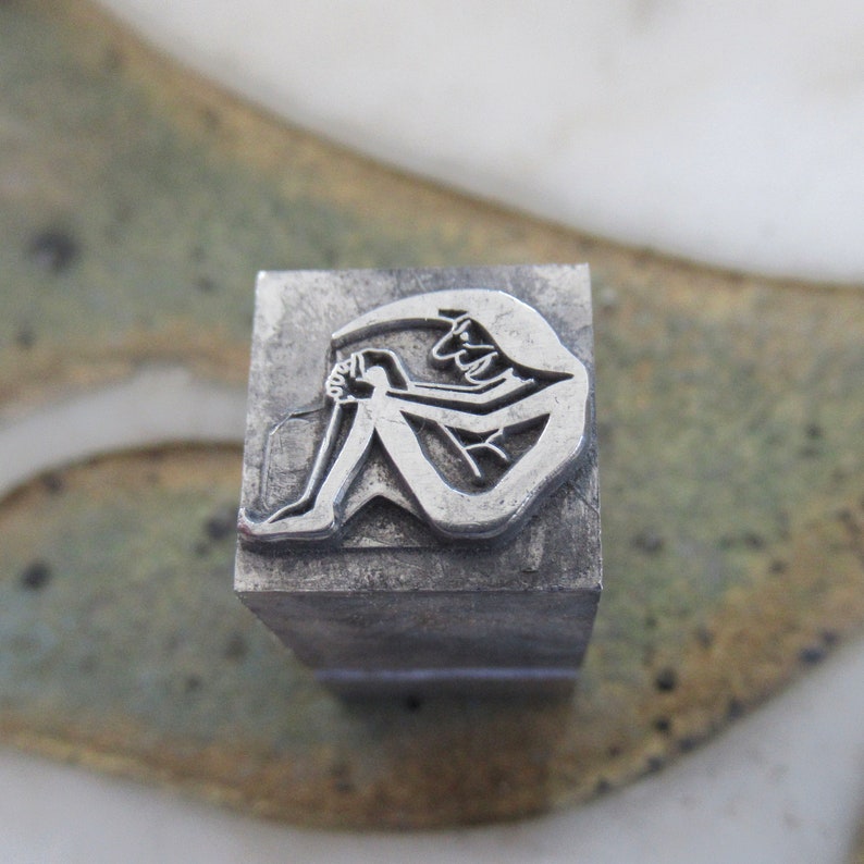 May include: A silver metal stamp with a design of a person sitting with their legs crossed and their arms wrapped around their legs.