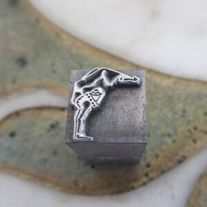 Op de afbeelding: Een metalen stempel met een ontwerp van een persoon die buigt, mogelijk een danser of gymnast. De stempel is zilverkleurig en heeft een getextureerd oppervlak.