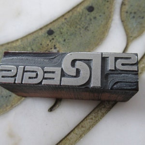 May include: A metal printing block with the word "SIGEL" embossed in raised letters. The block is made of a dark gray metal and has a wood base.