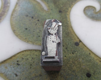May include: A vintage metal printing block featuring a black and white illustration of a person wearing a hat and uniform. The person is holding a stick and standing on a platform.