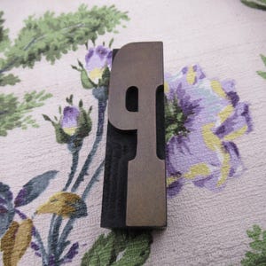 May include: A vintage metal number nine printing block. The number is in a serif font and has a dark gray color. The block is set against a floral patterned fabric with purple and green flowers.