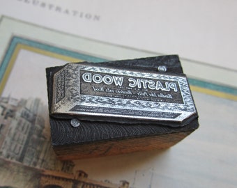 Puede incluir: Un bloque de impresión de madera vintage con una placa de metal plateado que dice "Plastic Wood" y "Handles like putty - Hardens into wood".