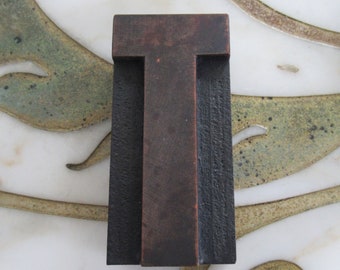 May include: A dark brown wooden letter T, with a slightly raised surface, is set against a white background with a decorative pattern.