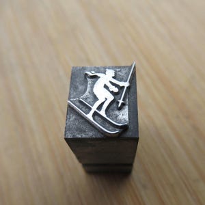 May include: A silver-colored metal stamp featuring a skier in motion, holding ski poles. The skier is depicted on skis, with a detailed outline. The stamp is mounted on a dark gray rectangular block, suitable for printing.