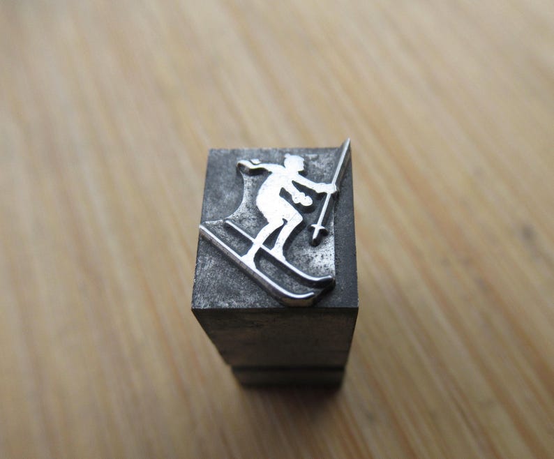 May include: A silver-colored metal stamp featuring a skier in motion, holding ski poles. The skier is depicted on skis, with a detailed outline. The stamp is mounted on a dark gray rectangular block, suitable for printing.