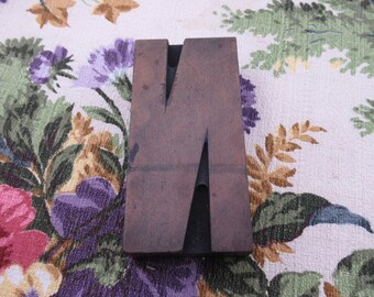 May include: A wooden letter N printing block. The block is dark brown with a lighter brown letter N carved into it. The block is sitting on a floral fabric background.