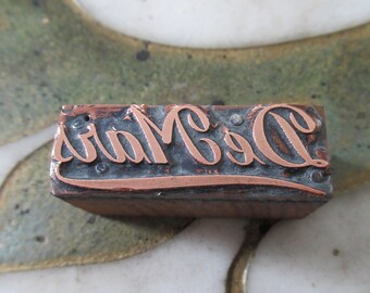 May include: A vintage wood printing block with the words "De Mars" in copper lettering.