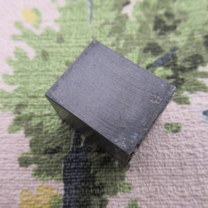 May include: A dark gray, square-shaped metal object. The cube has a slightly textured surface and appears to be made of a solid, heavy material. The edges are sharp, and the overall appearance is industrial.