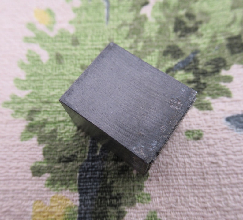 May include: A dark gray, square-shaped metal object. The cube has a slightly textured surface and appears to be made of a solid, heavy material. The edges are sharp, and the overall appearance is industrial.