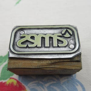 May include: A vintage metal stamp with the letters "sms" in a stylized font. The stamp has a rectangular shape with rounded corners and is mounted on a wooden block. The metal has a silver finish.