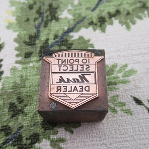 Può includere: Un blocco di stampa in metallo marrone con il testo "DEALER Nash SELECT 10 POINT" in lettere bianche. Il blocco ha un design a scudo color rame con una stella al centro.