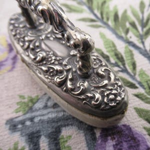 May include: An antique silver-coloured brush with an elaborate floral and scroll pattern. The brush features a curved handle and a rectangular base with a soft, light-coloured pad.