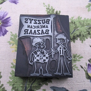 May include: A vintage printing block with a cartoon illustration of two figures holding a flag. The flag reads "Bussys American Bazaar" in reverse. The block is made of dark wood and metal.