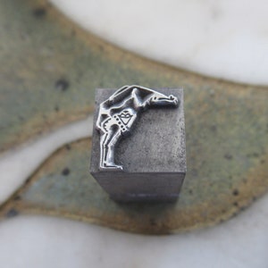 Op de afbeelding: Een metalen stempel met een ontwerp van een persoon die buigt met zijn armen boven zijn hoofd.
