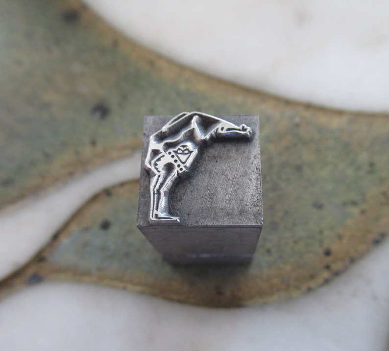 Op de afbeelding: Een metalen stempel met een ontwerp van een persoon die buigt met zijn armen boven zijn hoofd.