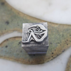 May include: A silver metal stamp with a design of a person sitting with their legs crossed. The stamp is rectangular and has a textured surface.
