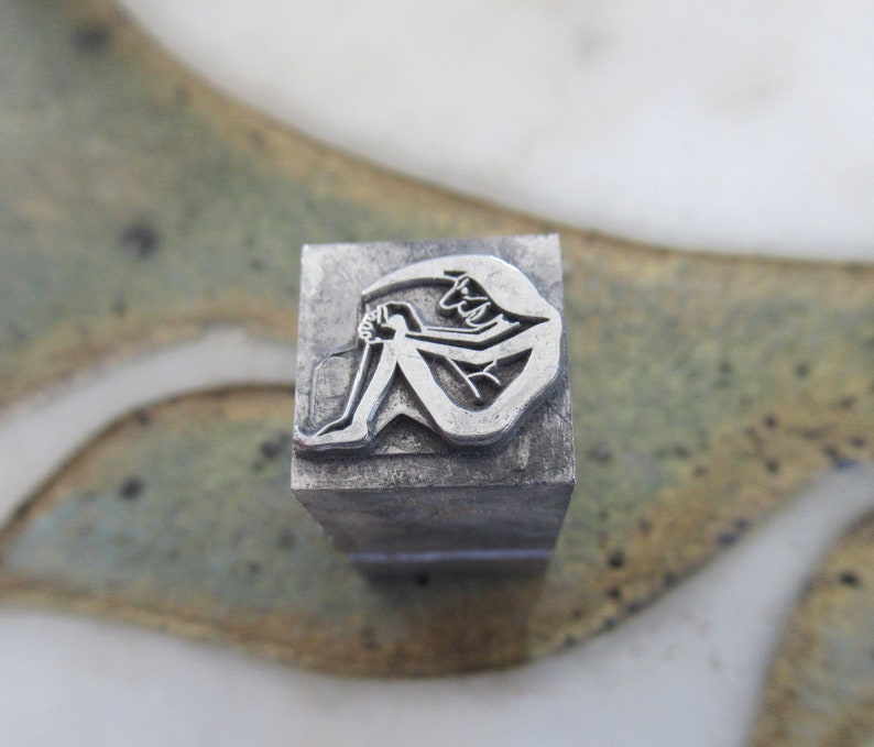 May include: A silver metal stamp with a design of a person sitting with their legs crossed. The stamp is rectangular and has a textured surface.