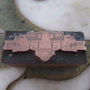 May include: A copper-colored metal stamp with the words "ЯR ОТЭМ ИG" engraved on it. The stamp is mounted on a black metal base.