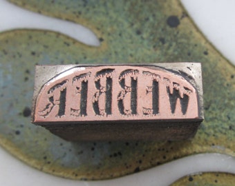 May include: A vintage metal letterpress printing block with the word "WIBBLE" in reverse. The letters are in a bold, serif font and are made of copper. The block has a rectangular shape with a silver metal frame.