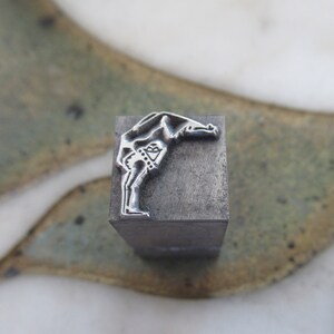 Op de afbeelding: Een metalen stempel met een ontwerp van een persoon die een handstand maakt. De stempel is grijs en heeft een rechthoekige vorm.