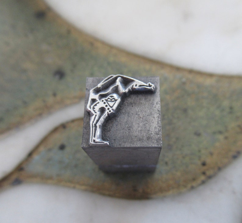 Op de afbeelding: Een metalen stempel met een ontwerp van een persoon die een handstand maakt. De stempel is grijs en heeft een rechthoekige vorm.