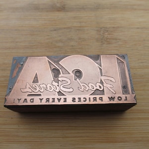 May include: A rectangular copper printing block with the text "Food Stores" and "ISA" in a vintage font. The block also includes the text "LOW PRICES EVERY DAY!" in reverse. The block is set against a wooden background.