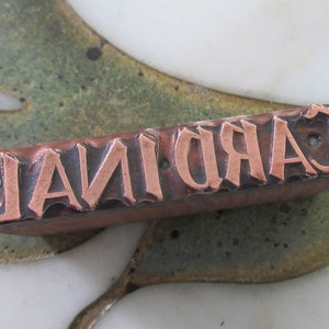 Op de afbeelding: Een vintage metalen drukblok met het woord "CARDINAL" in koper gegraveerd.
