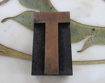 May include: A vintage metal letterpress block with the letter "T" in a copper color. The block is surrounded by a black frame.