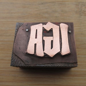 May include: A rectangular, copper-colored stamp with the letters "AGI" in a stylized font. The letters are a lighter copper color and are raised above the surface. Two small, silver-colored rivets are visible on the stamp.