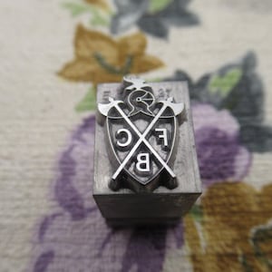 May include: A metal stamp with a shield design featuring the letters "C", "E", and "B" crossed by two axes. The stamp is set on a rectangular block and is likely used for imprinting designs.