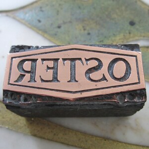 Op de afbeelding: Een vintage drukblok met een koperkleurige plaat waarop "OSTER" in een decoratief lettertype staat.