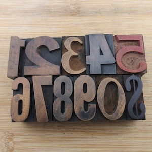 May include: A collection of vintage wooden printing press numbers in various colors, including brown, gray, and red. The numbers are arranged in two rows on a light wooden surface.