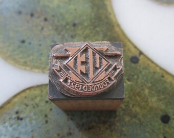 May include: A close-up of a metal stamp with a copper-coloured design. The design features a diamond shape with the letters "EU" inside and the words "Founded 1931" around the outside.