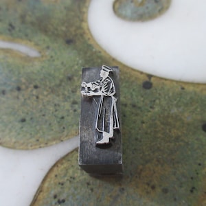 Może przedstawiać: Metalowy blok drukarski z wypukłym obrazem osoby w mundurze trzymającej tacę. Osoba stoi na platformie, a obraz jest otoczony obramowaniem.