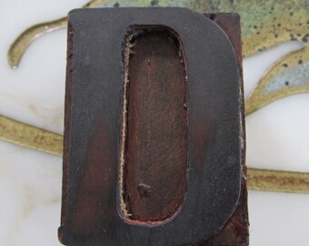 May include: A dark brown wooden letter D with a black outline. The letter is raised and has a slightly worn look.