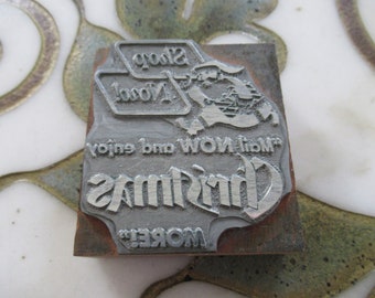 Könnte beinhalten: Ein silberner Metalldruckblock mit den Worten "More" "Christmas" "Mom and Dad enjoy" und "Shop Now!" Der Block zeigt ein stilisiertes Bild einer Person mit Hut und Tasche.