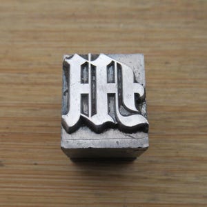 May include: A vintage metal letterpress block featuring the letter "M" in a decorative, ornate font. The block is silver-colored and sits on a wooden surface. The letter is raised and detailed, showcasing a classic printing style.
