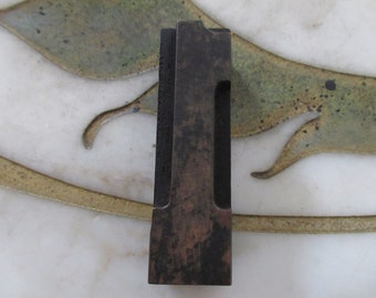 Può includere: Un blocco di stampa in legno marrone scuro, di forma rettangolare, con una sezione scavata. Il blocco mostra segni di usura e invecchiamento. Poggia su una superficie bianca con un motivo decorativo oro e verde, creando contrasto.